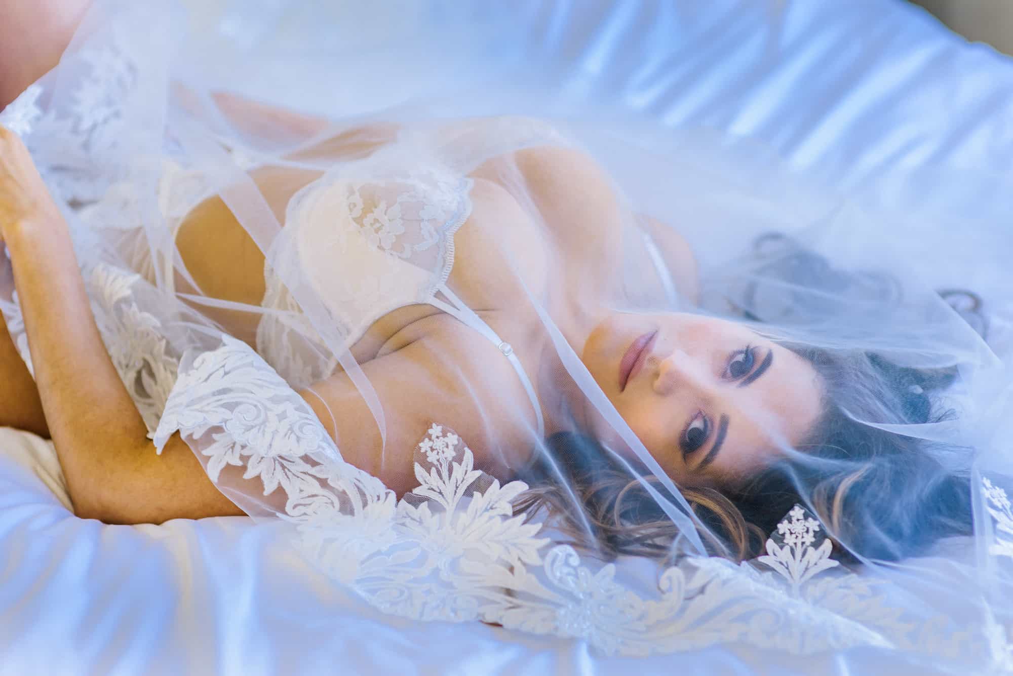 bride in her bridal lingerie laying down on the bed covered by her veil