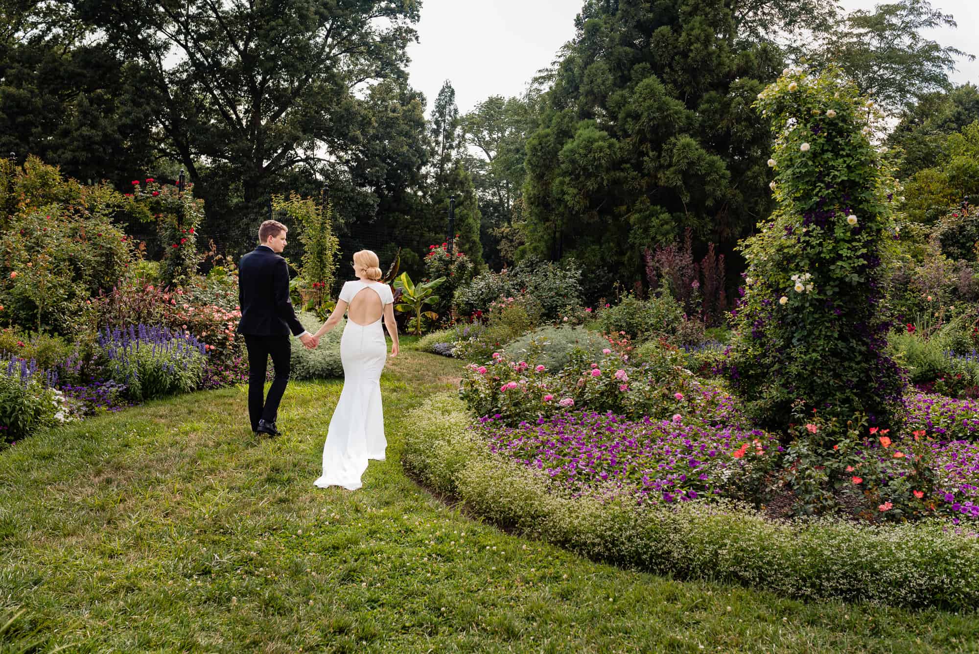 bride and groom in a beautiful field of flowers walking while holding hands at their Morris Arboretum Wedding