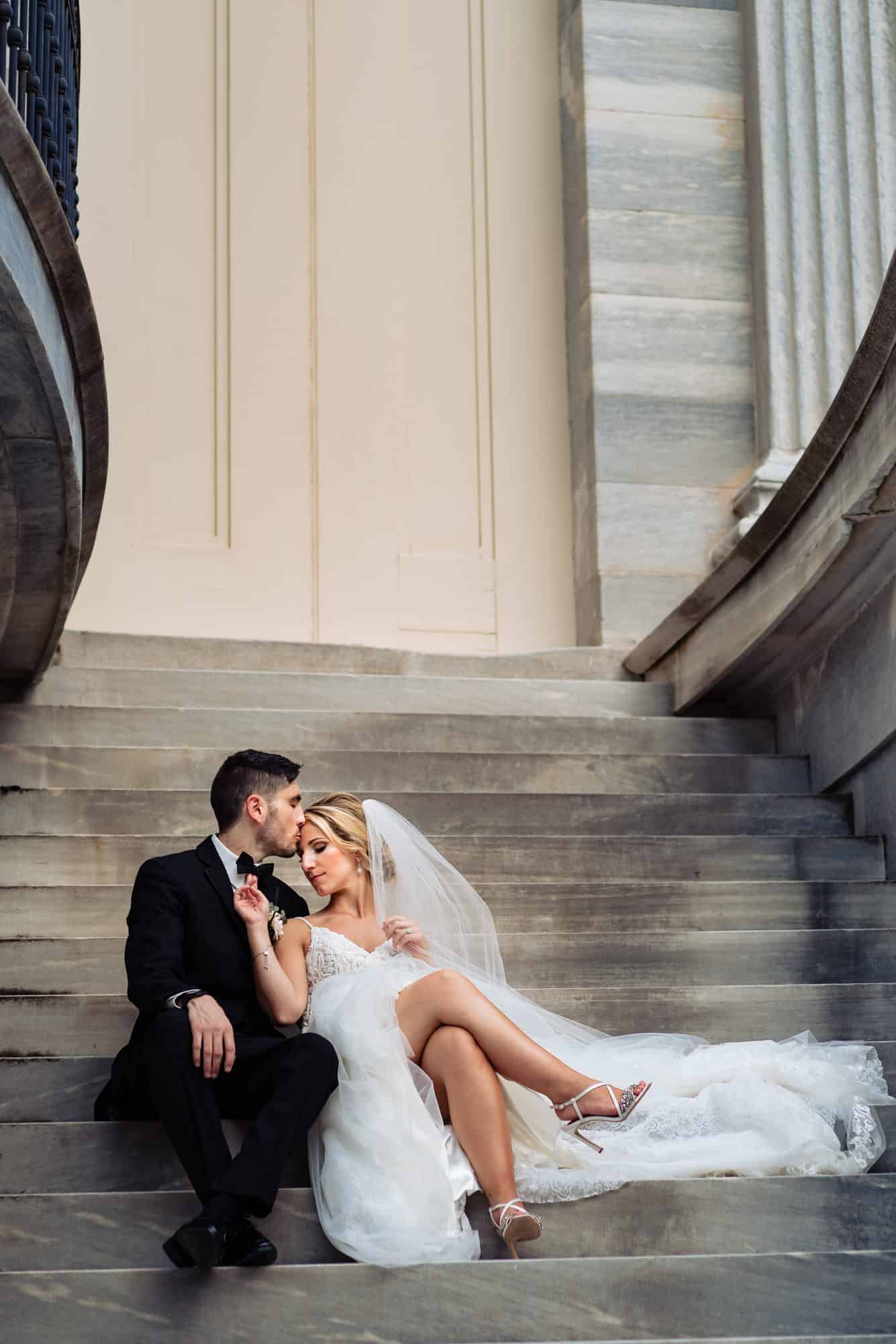 groom is kissing her bride's forehead while sitting at the stairs