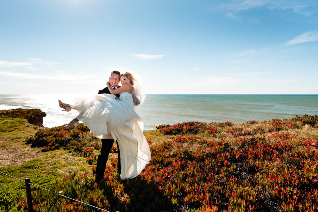 groom carrying bride on a cliff next to the ocean at their Ritz Carlton Half Moon Bay Wedding