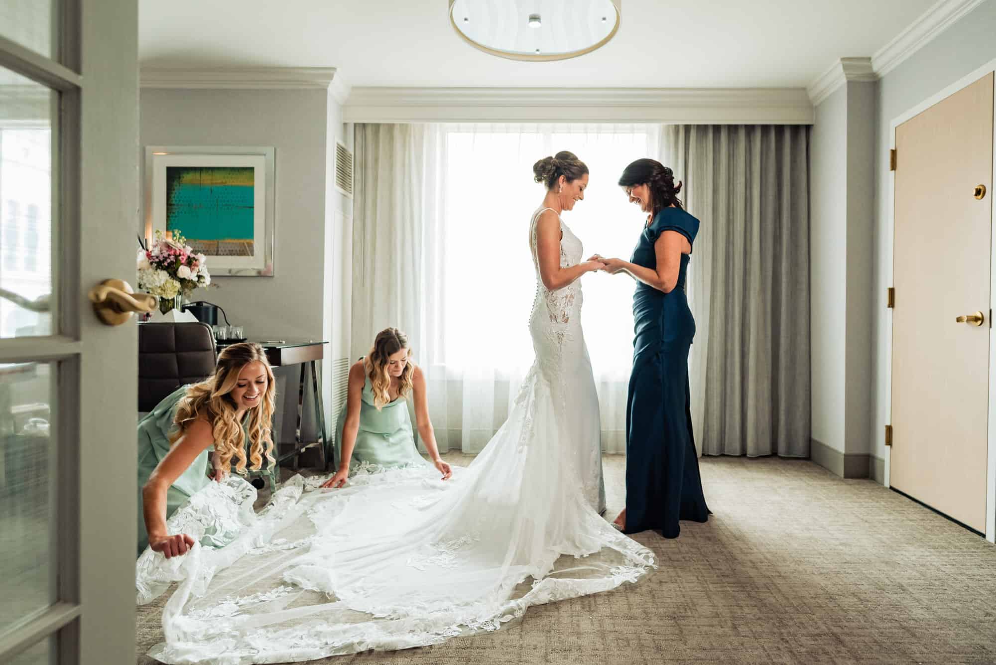 bride and her mother holding hands while the bridesmaids fix her gown