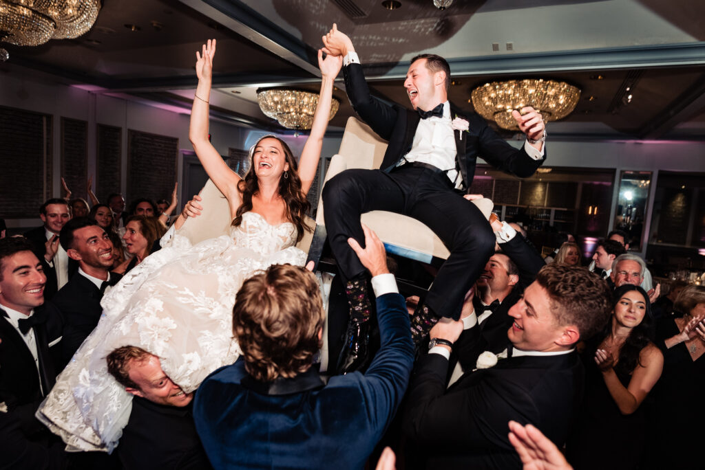 bride and groom are seated on chairs and lifted into the air in a celebratory dance at the reception called the hora