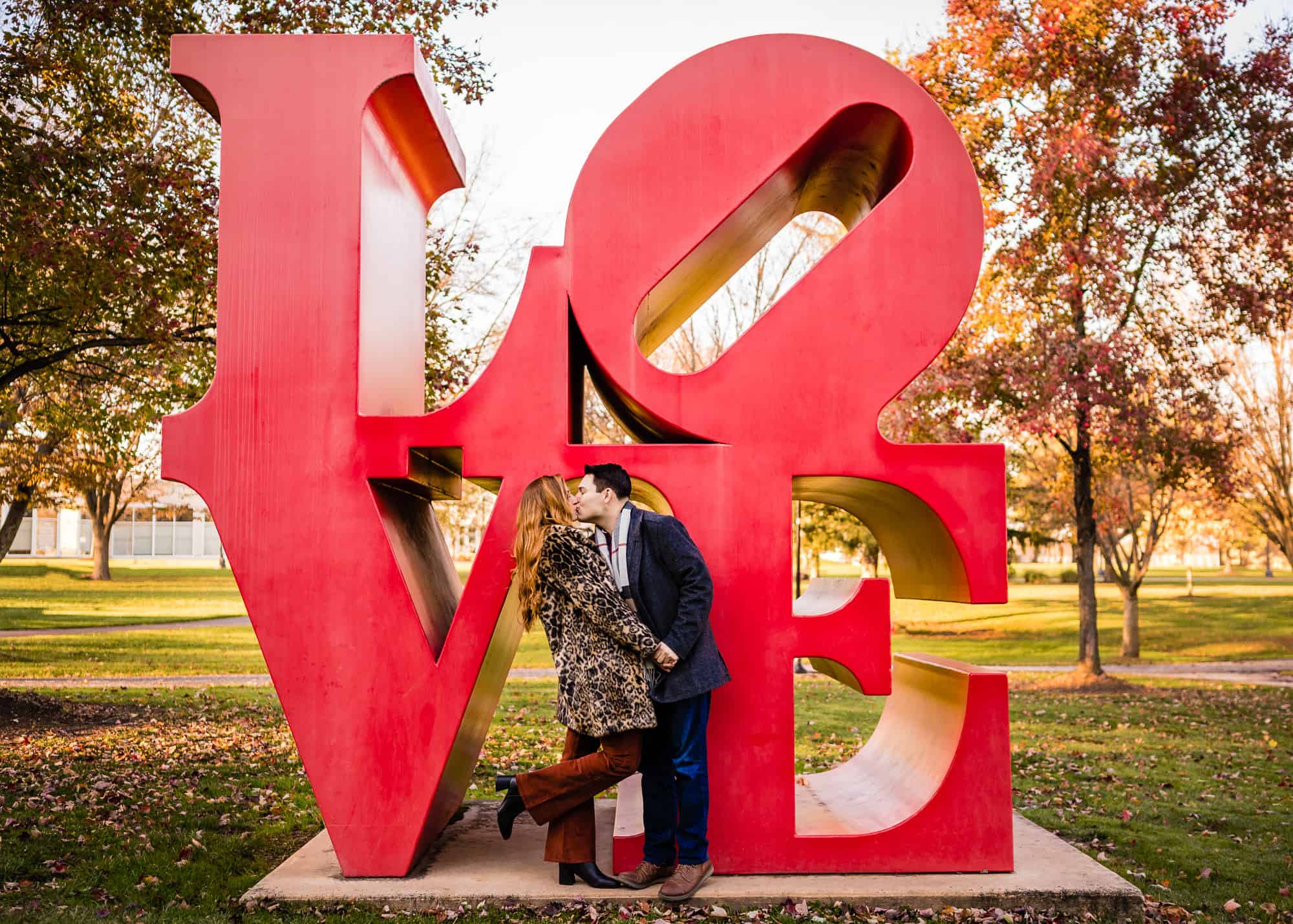 couple shares a kiss with the love sculpture designed by Robert Indiana located on the grounds of Ursinus College during their engagement photoshoot