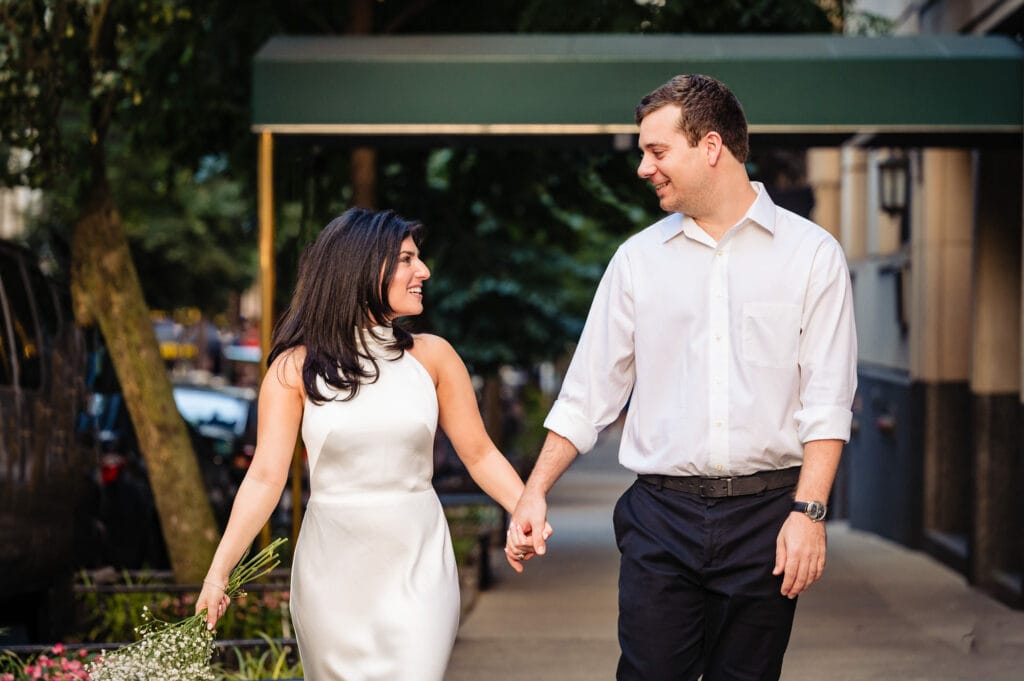 man and woman holding hands and looking at eachother walking on the street while the woman holds flowers