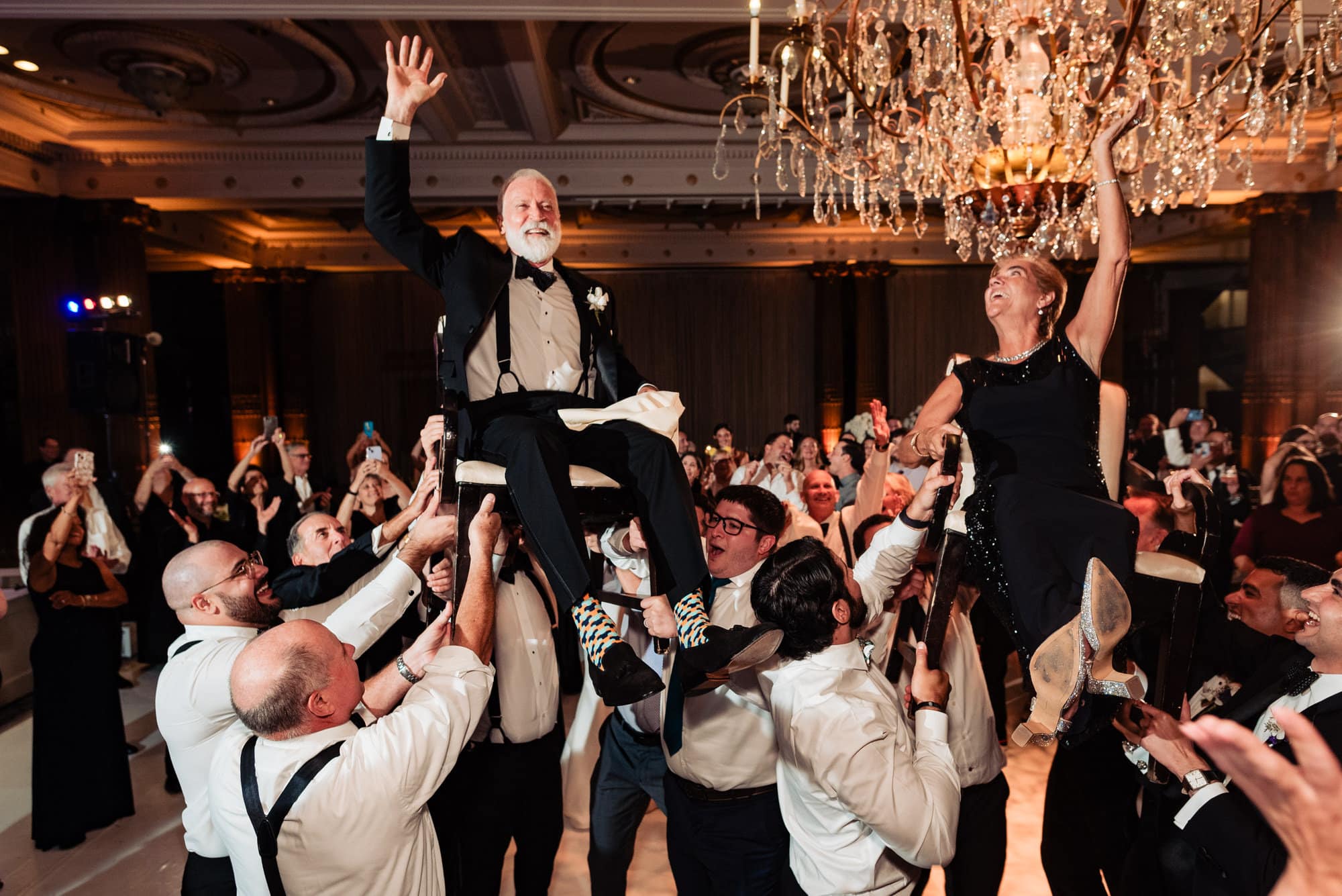 the groom's parents is carried on a chair while surrounded by guests