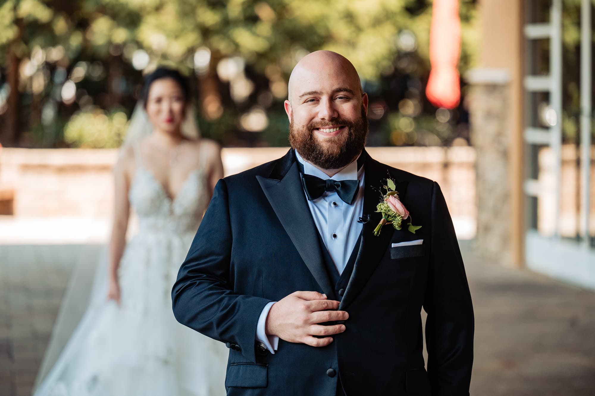 groom smiles for the camera while his bride stands behind him for their first look