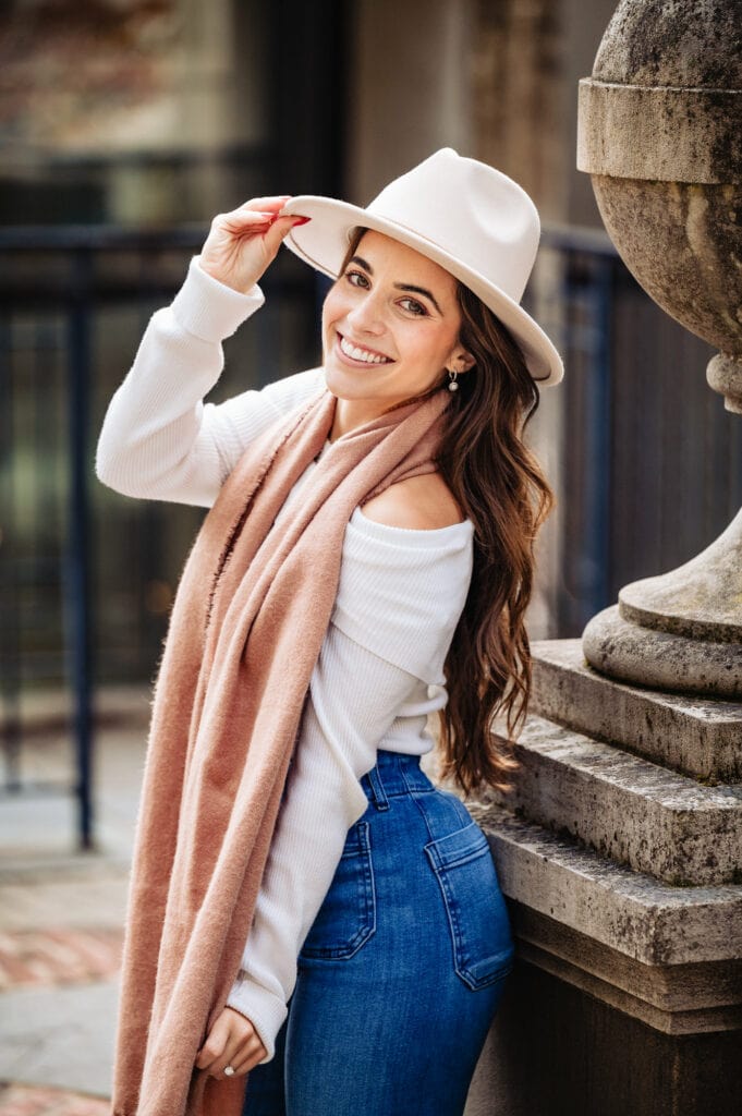 Woman in a white long sleeve top and cap, donning a brown shawl around her neck, radiates a beautiful smile during her engagement photoshoot.