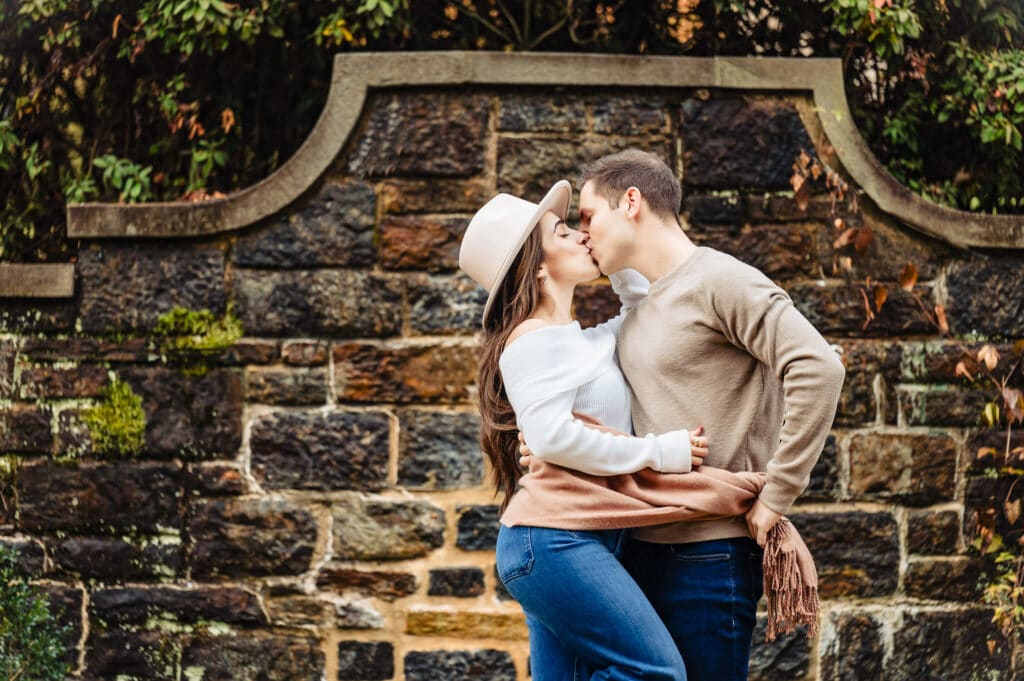 The couple kisses as the man wraps a scarf around the woman's waist, pulling her closer against the beautiful backdrop of the fall scenery.