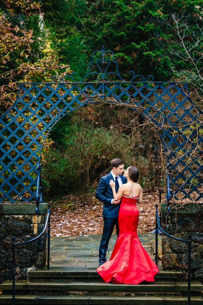 An engaged couple sharing a romantic kiss at a garden gate entrance, intricate wrought iron designwork pillar framing them. Morning light streams through the pillars encircling their private moment during their creative fall engagement photo shoot.