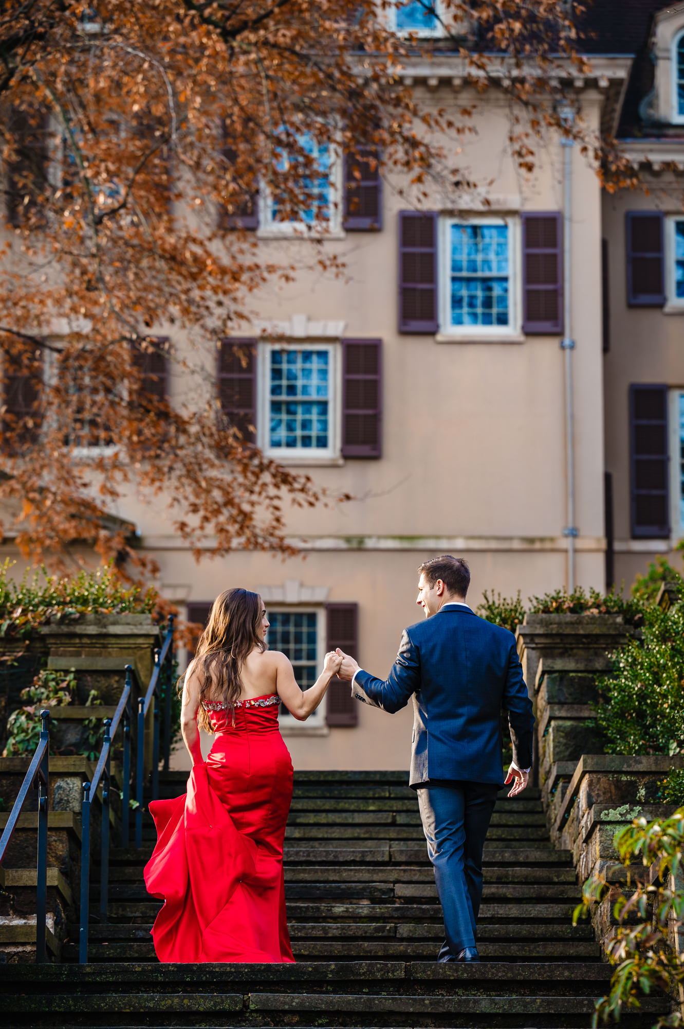 An engaged couple walks on stone steps lined with colorful fallen leaves leading to the historic mansion visible in the background of their intimate photo.
