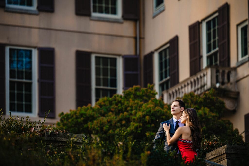 An elegantly dressed engaged couple turns away from each other, striking a dramatic pose framed by red and orange autumn foliage. The woman in a dress looks off in one direction as her tuxedo-clad fiancé gazes off in the other, his arm around her bare waist.