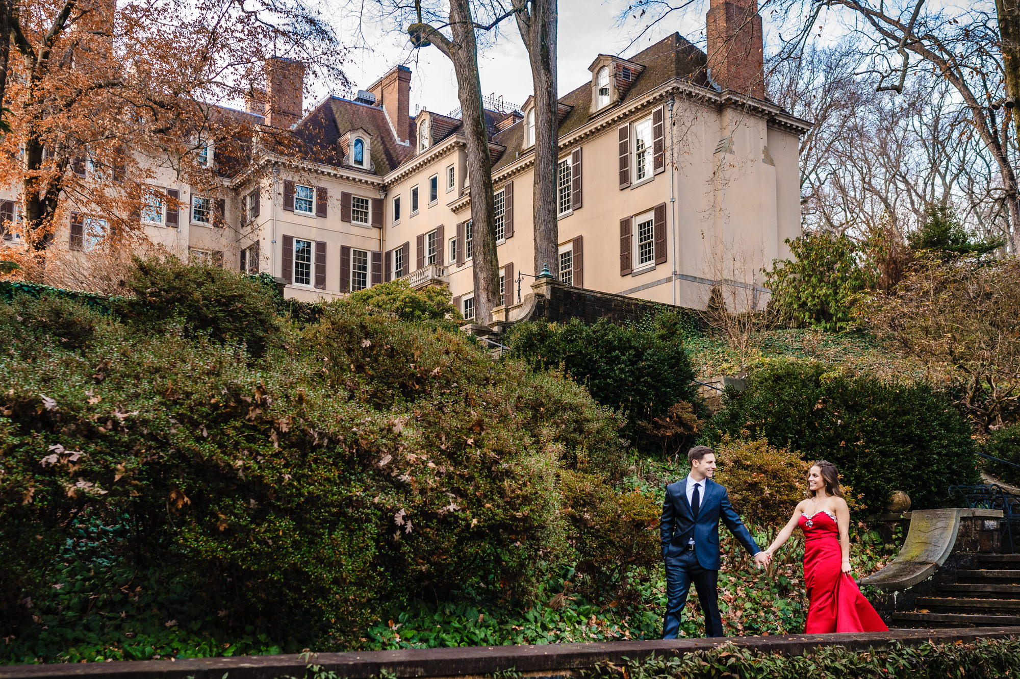An engaged couple walk while holding hands affectionately. Stone steps lined with colorful fallen leaves lead to the mansion visible in the background of their intimate photo.