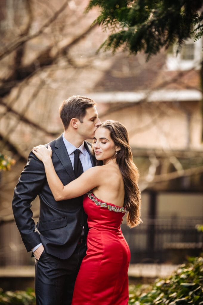 An engaged couple cherishing an intimate moment, tenderly locked in a warm embrace amidst the vibrant fall foliage. The man affectionately places a gentle kiss upon his smiling fiancée's forehead as she clutches him, eyes closed.