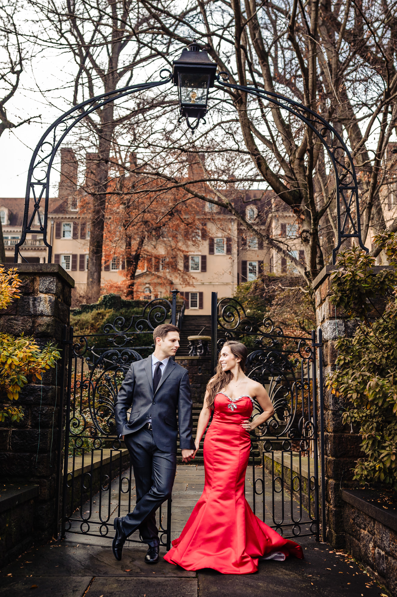 An engaged couple stands affectionately hand-in-hand at an ornate garden gateway, smiling brightly at the camera both glowing with happiness. An intricate iron gate and stone pillars frame the joyful couple, with a tree-lined path and grand historic mansion visible in the background.