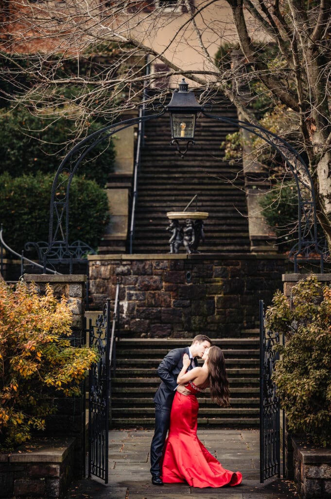 An engaged couple sharing a romantic kiss at a garden gate entrance, intricate wrought iron designwork and stone pillars framing them. The man cradles his smiling fiancée's face affectionately in his hands as she wraps her arms around his shoulders. Stone steps lined with colorful fallen leaves lead to the historic mansion visible in the background of their intimate photo.