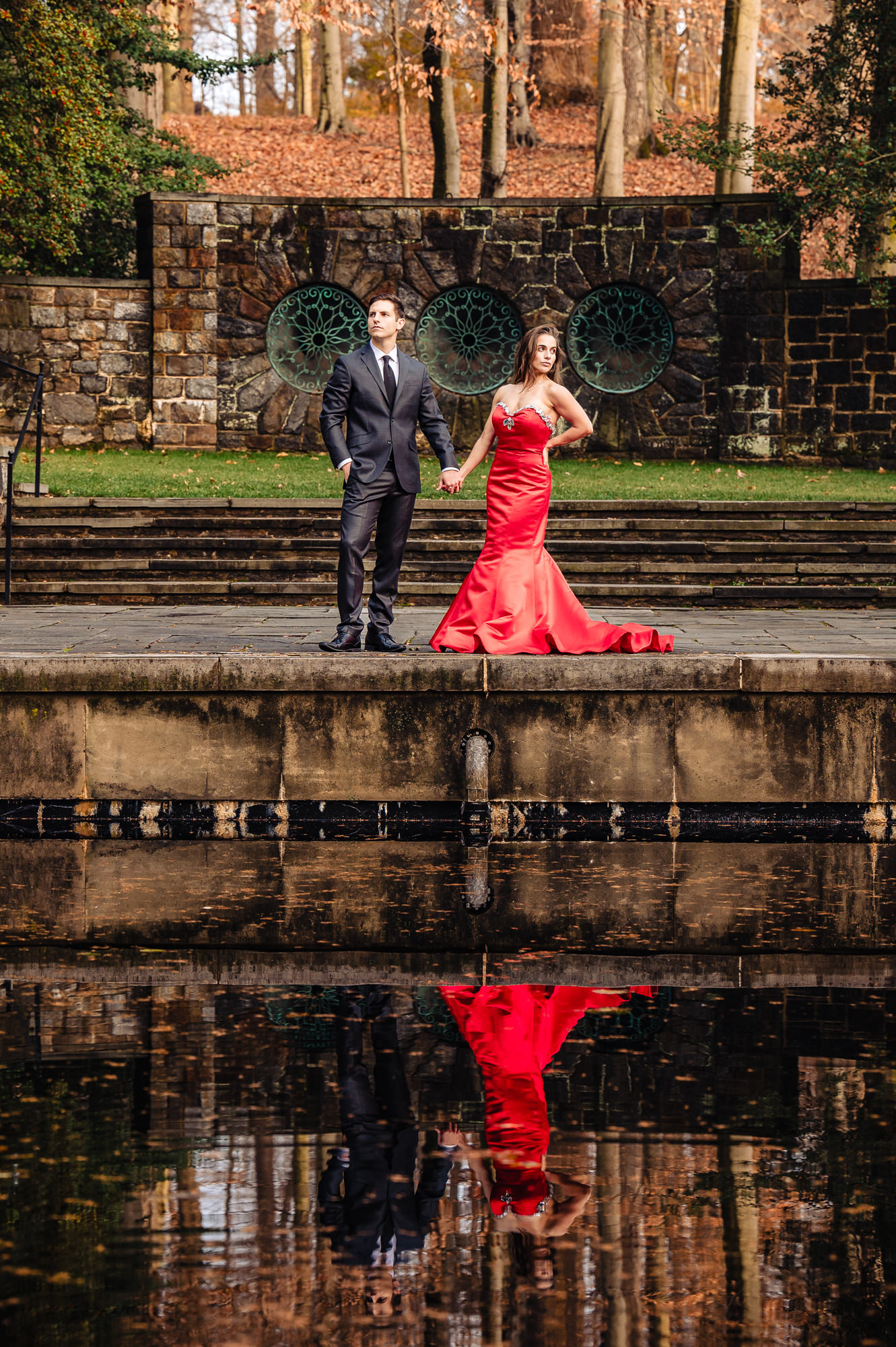 An elegantly dressed engaged couple turns away from each other, striking a dramatic pose in front of a stone pool framed by red and orange autumn foliage. The woman in a red gown looks off in one direction as her suit-clad fiancé gazes off in the other while holding each other's arms.
