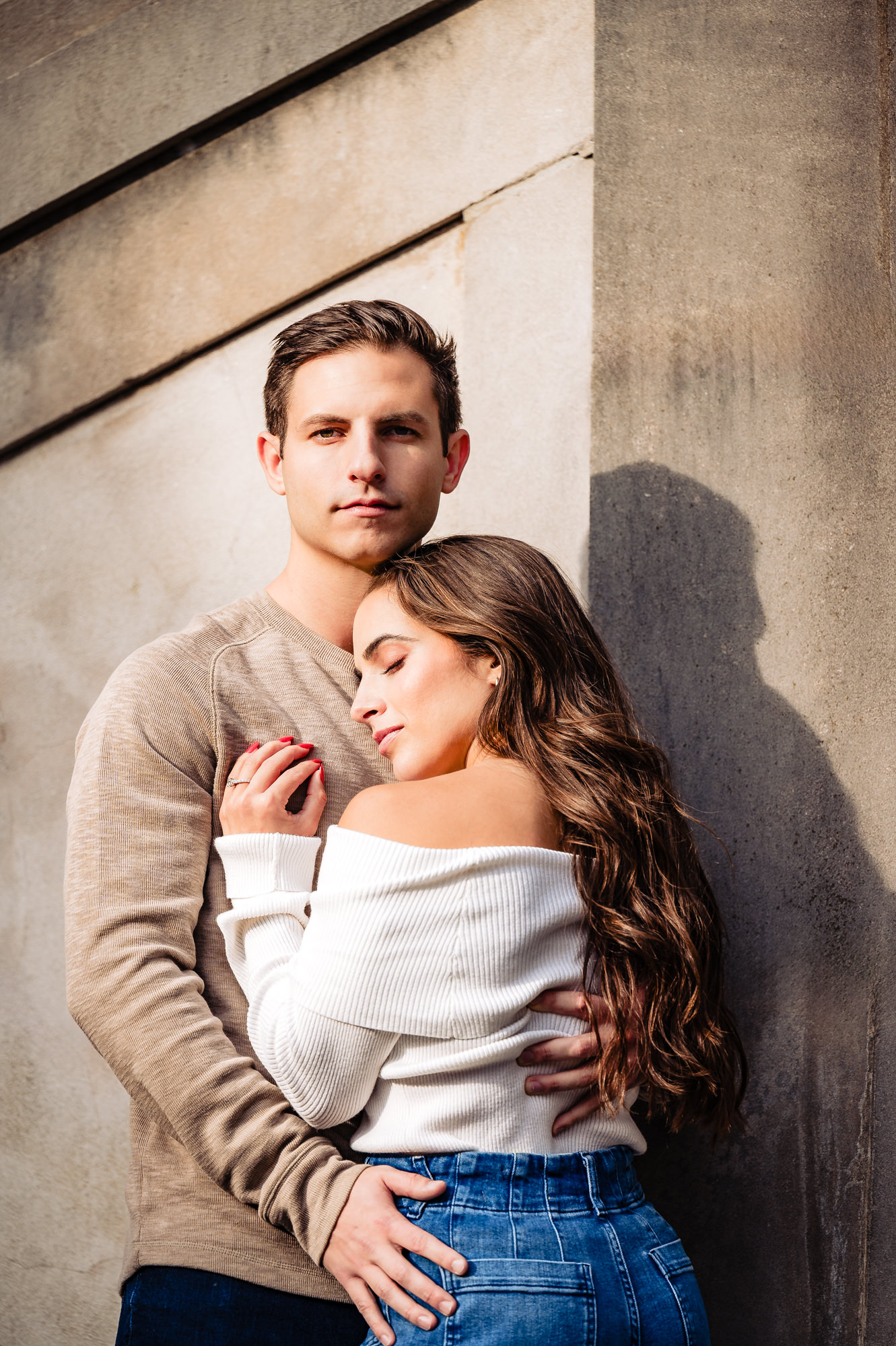 An engaged couple cherishing an intimate moment, tenderly locked in a warm embrace against the backdrop of a striking concrete structure.