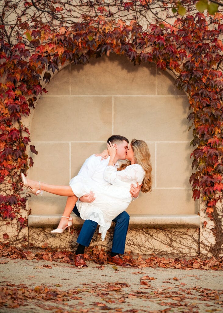 A young man and woman are sitting together on a stone bench, surrounded by the vibrant red and orange leaves of maple trees in autumn. The woman is tucked closely into the man's arm, both smiling softly as a light breeze rustles the fallen leaves scattered across the ground.