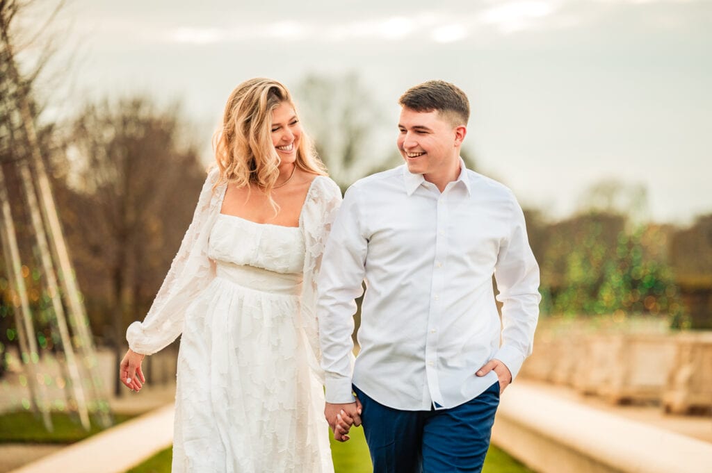 an engaged couple embraces amidst the scenic beauty of Longwood Gardens. Smiling joyfully, they gaze into each other's eyes, barely noticing the camera capturing the memorable moment.