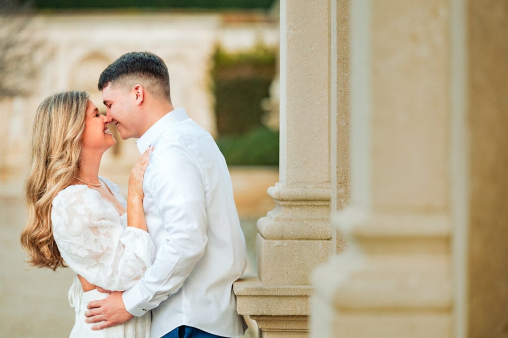 The beaming couple walk hand-in-hand down a garden path lined with vibrant fall foliage in oranges, reds and yellows. Turning towards one another, they smile brightly, overflowing with happiness on the occasion of their engagement and upcoming marriage.