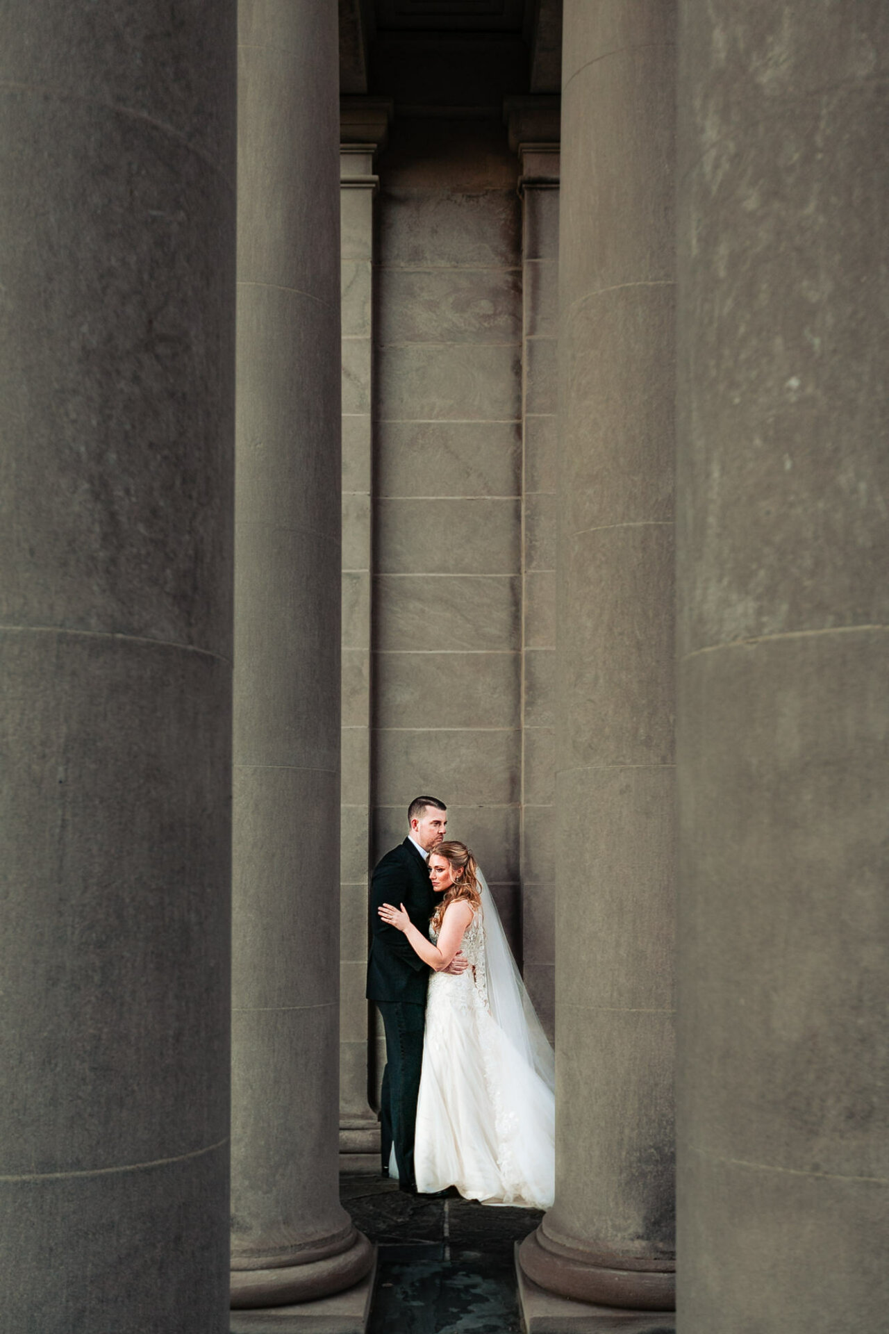 A newly married couple poses affectionately together in an urban setting, standing between massive concrete columns that frame them dramatically.