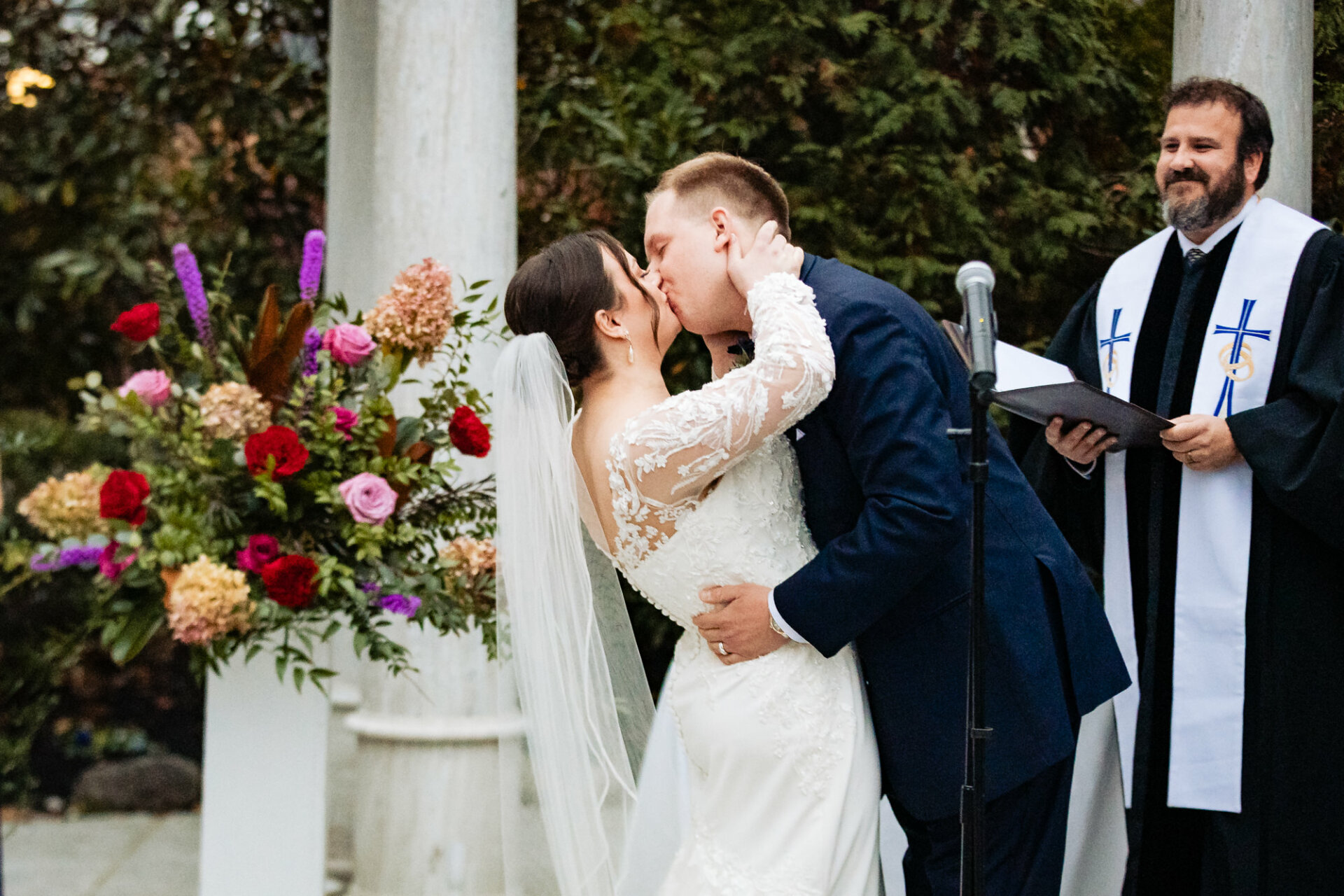 Captivating moment as the newlyweds share a tender kiss, framed by the smiling priest. This heartwarming scene encapsulates the joy and blessings bestowed upon the couple as they embark on their journey together in the sanctity of marriage