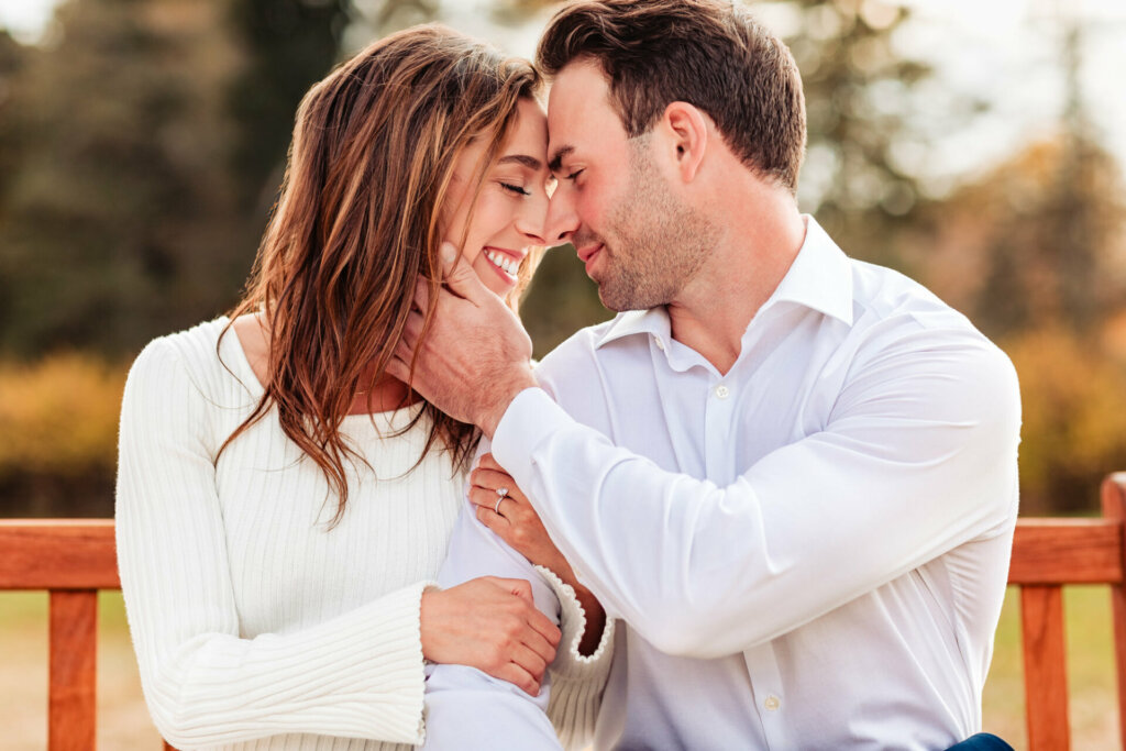 Serene moment captured as a man and woman sit on a bench, leaning their foreheads on each other in a tender embrace. The woman holds the man's arm, while he reciprocates by gently holding her cheek. The scene radiates intimacy and connection in a peaceful setting.