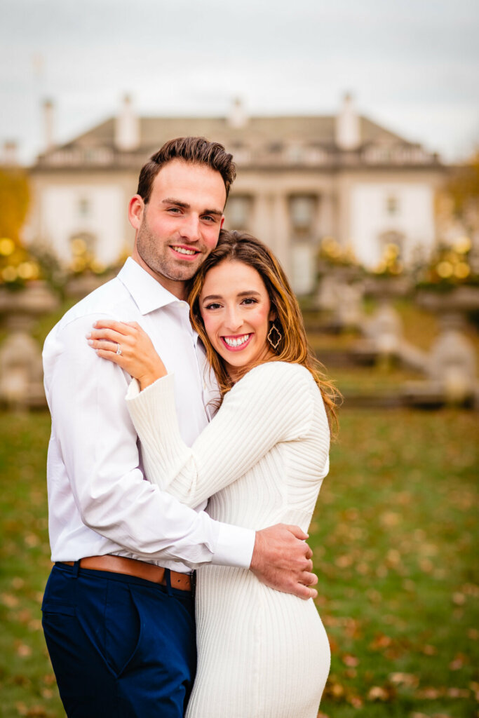 Enchanting embrace of a man and woman, both smiling at the camera, with the stunning Nemours Estate in the background. The joyous moment and picturesque setting come together, creating a timeless and beautiful image of love against the backdrop of the elegant estate.