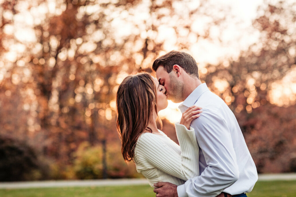 Romantic scene of a man and woman sharing a kiss, bathed in warm sunlight amidst a backdrop of the trees with orange leaves. The golden glow adds a touch of enchantment to the moment, creating a captivating and intimate image of love in nature.