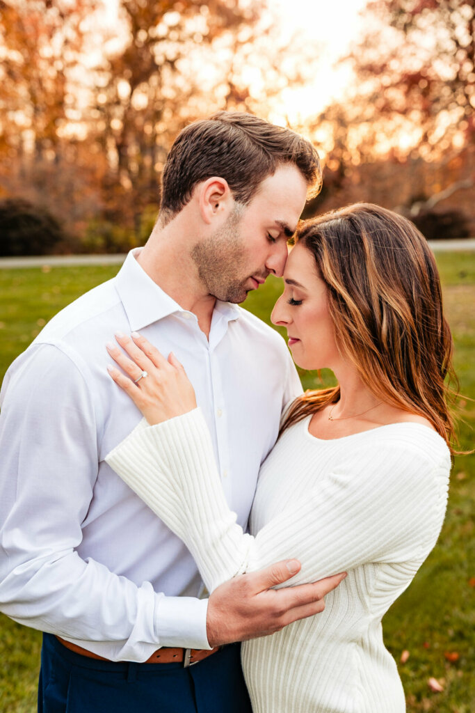 A serene scene featuring a woman in a long-sleeved white dress and a man in a long-sleeved button-up, facing each other and holding one another with their foreheads touching. The backdrop showcases orange trees and a lush green field, creating a picturesque and romantic setting for this intimate moment.
