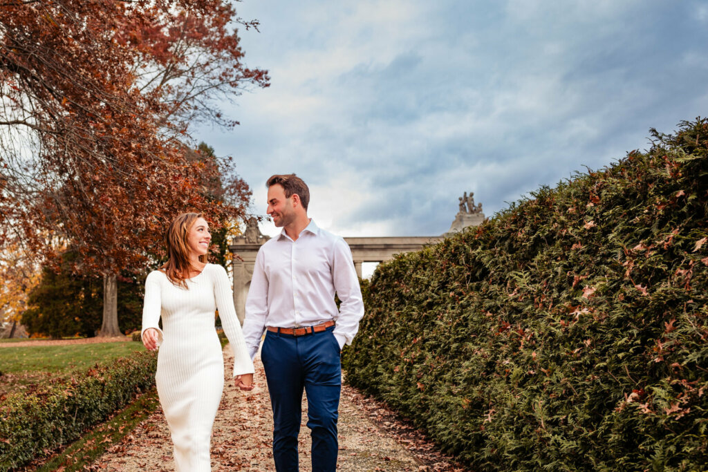 Enchanting scene as a man and woman walk hand in hand along a pathway, flanked by a creatively styled bush resembling a wall on one side. Their shared journey is beautifully framed by the greenery, creating a charming and intimate moment.