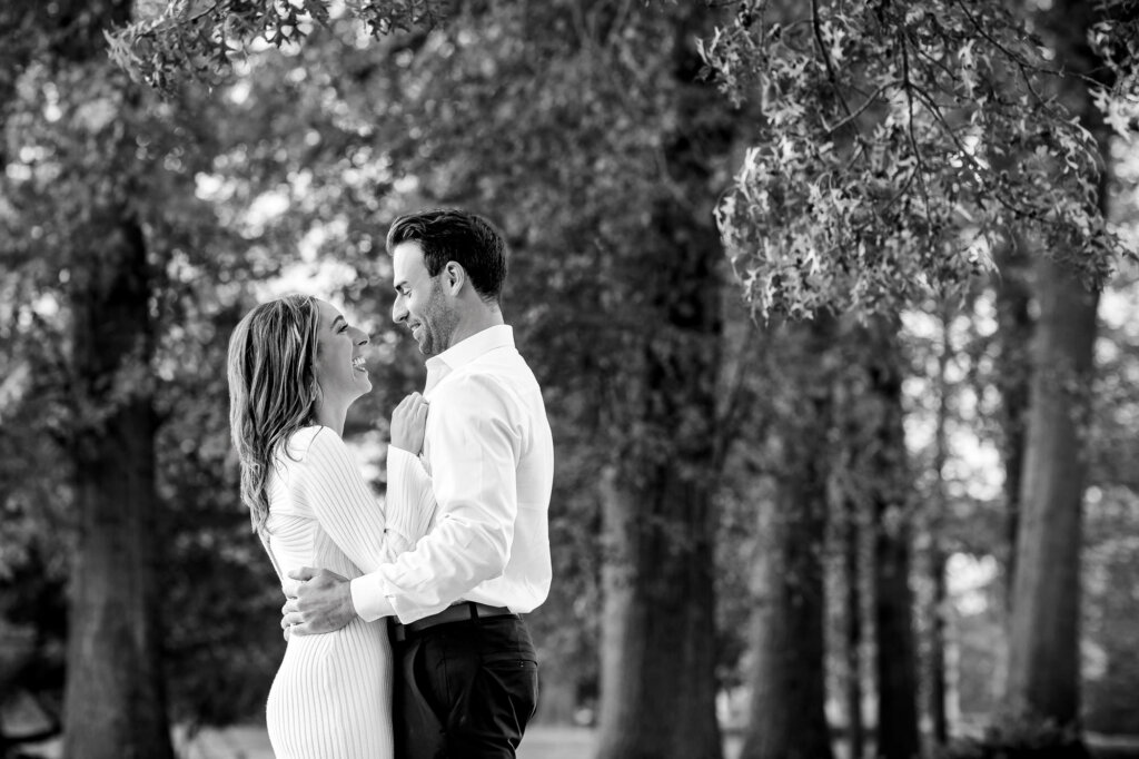 Timeless black and white photograph capturing the couple facing each other, with the man's hand placed gently on the woman's hips. Both share genuine laughter, creating a candid and joyful moment frozen in monochrome elegance.