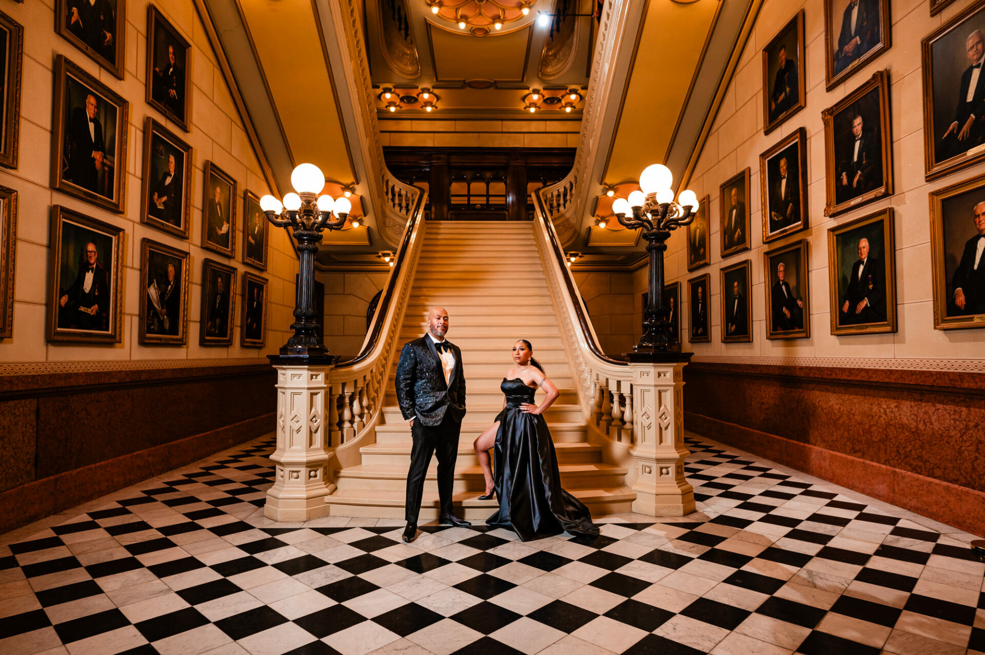 In an engagement photoshoot set against the backdrop of ancient architecture and expansive grand staircases at One North Broad, a fashionably attired couple is elegantly portrayed. The woman is clad in an off-the-shoulder, figure-hugging black dress with sheer sleeves, while her partner is dressed in a sleek black suit. They are surrounded by portraits of the Free Mason founders, with a majestic staircase providing a striking visual element behind them.