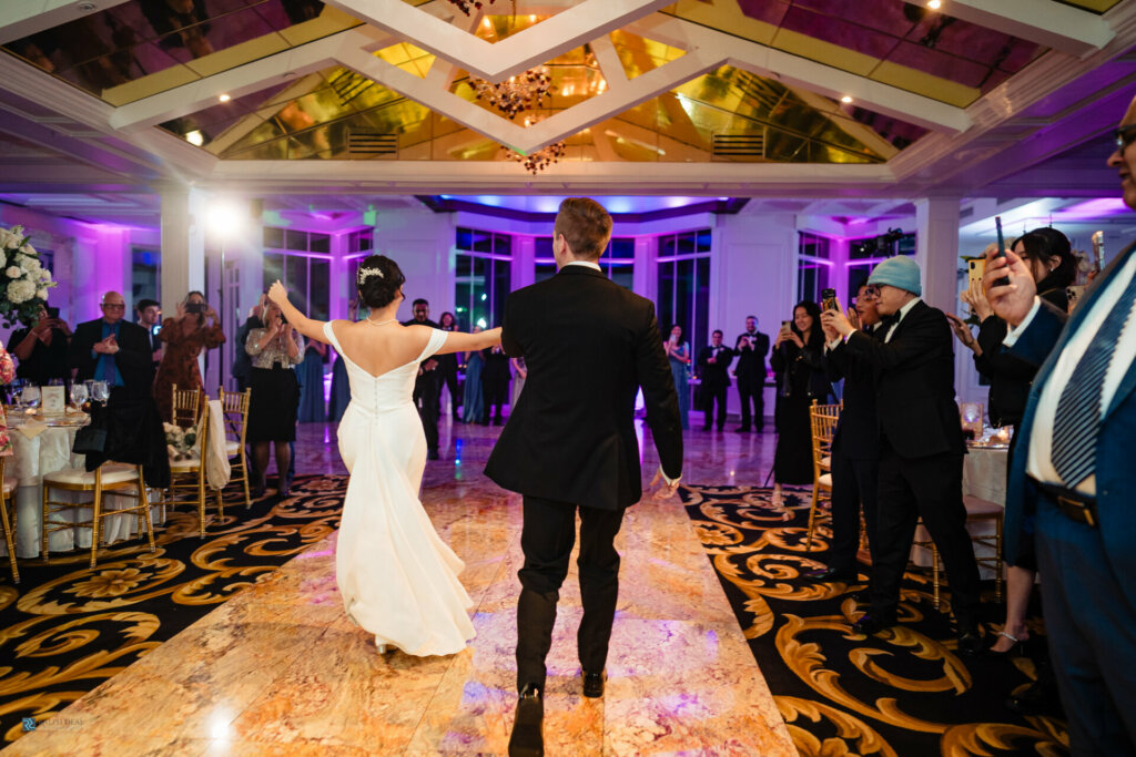 Captivating back shot capturing the groom and bride walking into the wedding reception, greeted by the enthusiastic happiness of the welcoming guests. The scene exudes warmth and joy as the couple enters the celebration, surrounded by the heartfelt cheers of their loved ones.