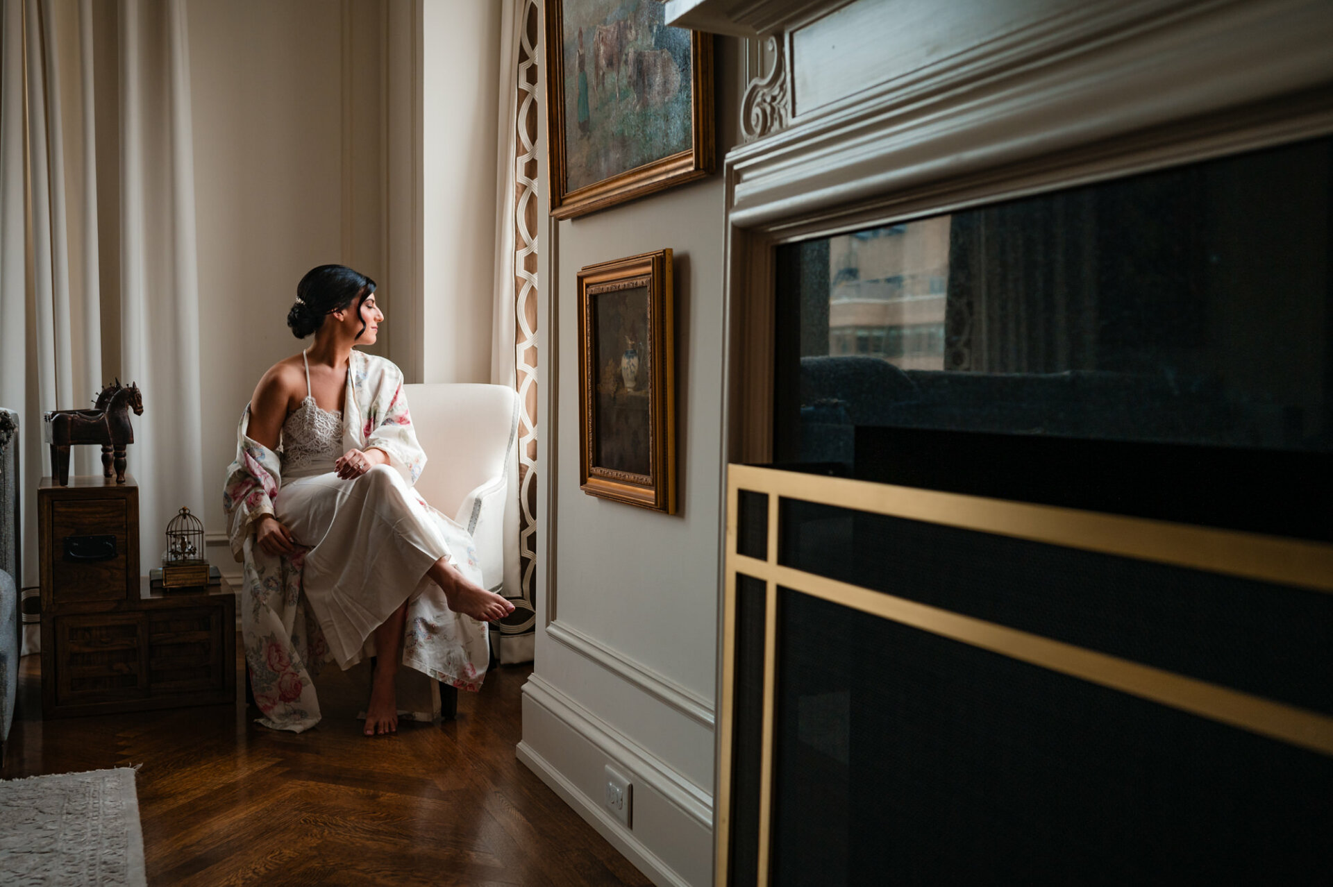 The bride, draped in her robe, sits gracefully in a white chair, gazing thoughtfully out of the window, capturing a serene moment of contemplation before her wedding.