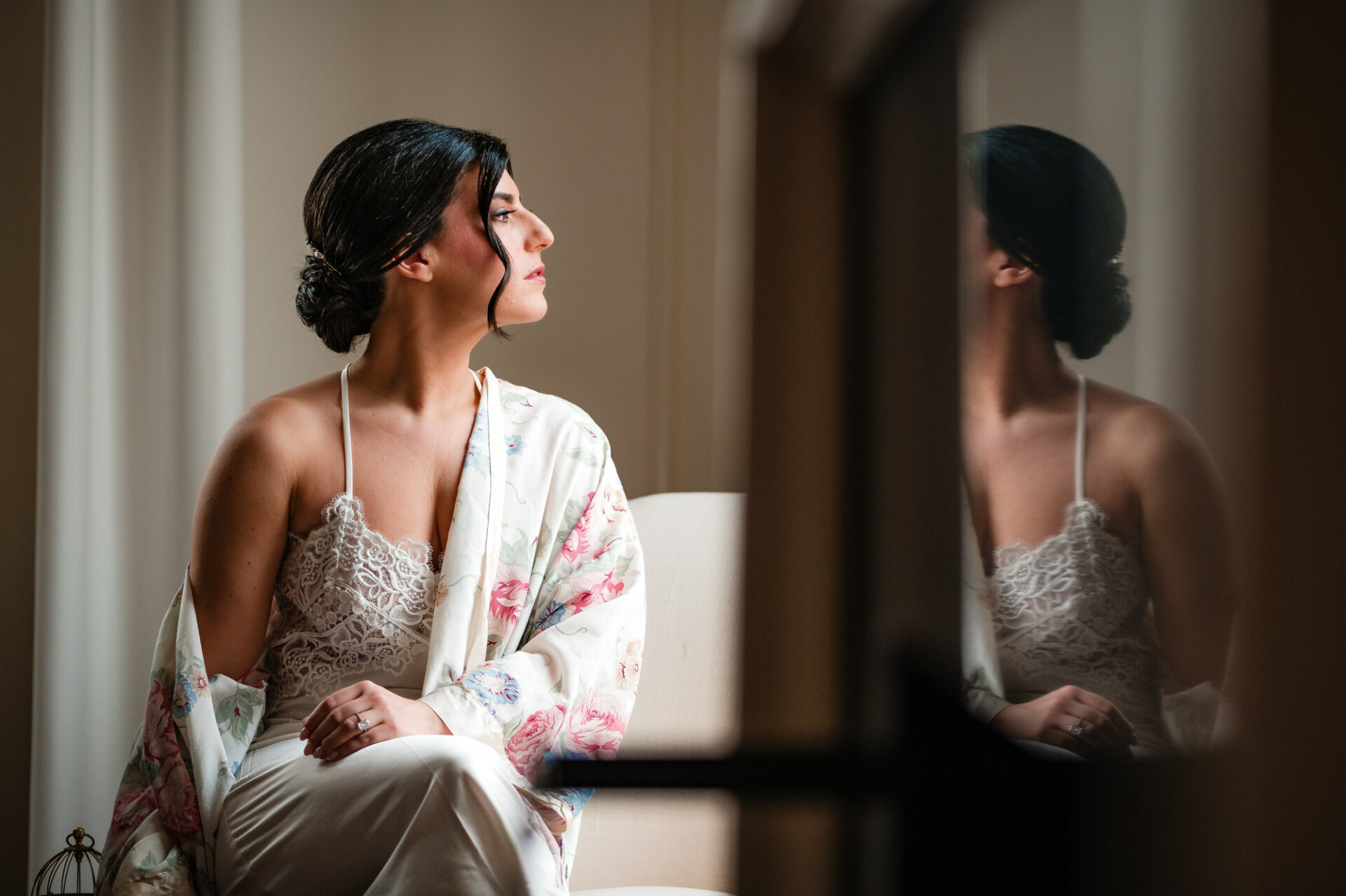 The bride, clad in her robe, sits gracefully in a white chair, her gaze directed out the window, perhaps pondering the moments ahead of her wedding day.