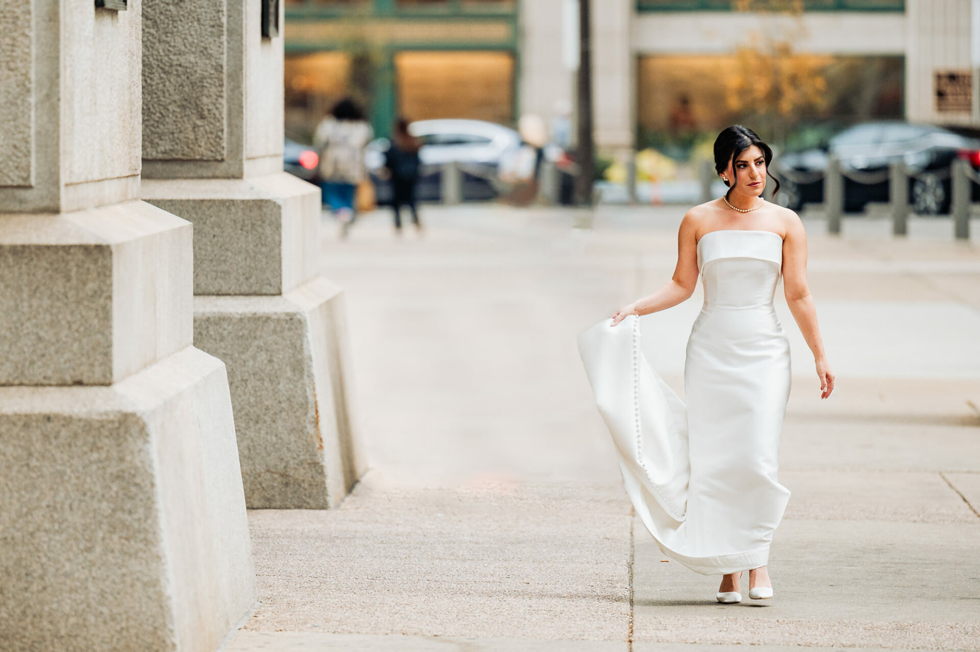 The bride gracefully walks along the street, carrying her train with elegance and confidence, a vision of beauty on her way to the ceremony.