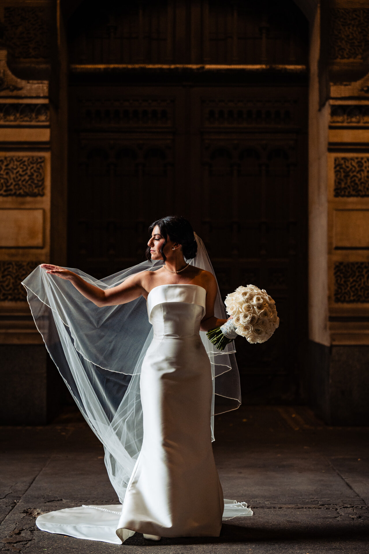 In a captivating solo bridal portrait, the bride gazes sideways with elegance and grace. Her delicate fingers gently touch her veil, while she holds her bouquet, exuding timeless beauty and anticipation on her wedding day.