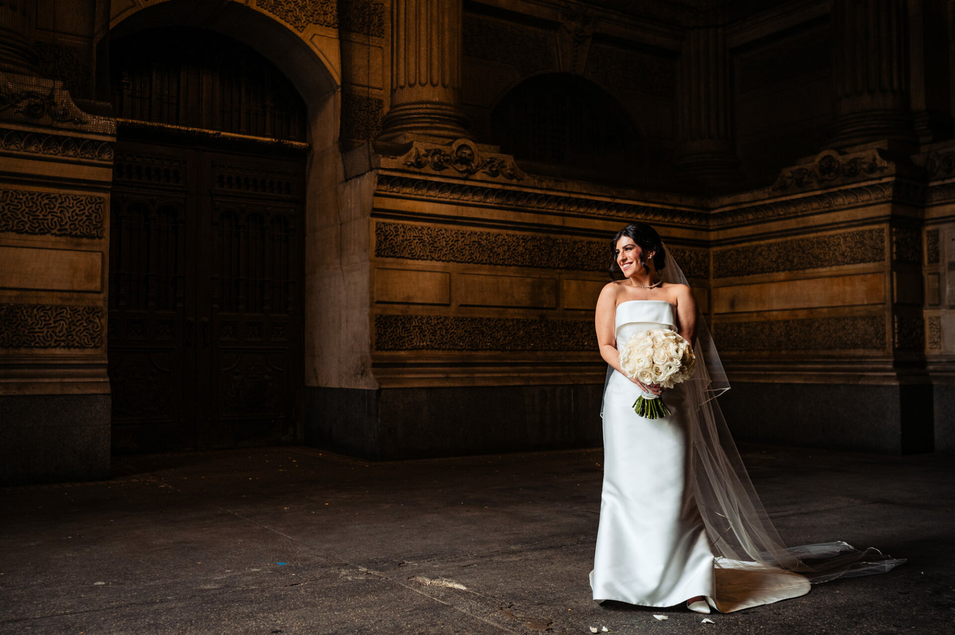 In a radiant bridal portrait, the bride beams with joy as her veil flows gracefully behind her. Her smile illuminates the frame, capturing the essence of happiness and excitement on her wedding day.