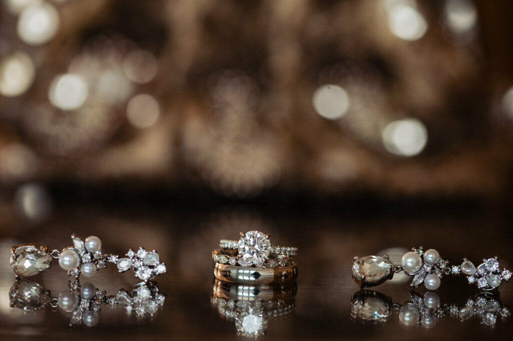 Showcasing bridal accessories, this still life photograph depicts objects laying elegantly. At the center rests an extravagant diamond eternity band engagement ring atop a lacy handkerchief. Surrounding it are two dangling diamond drop earrings that sparkle brilliantly.