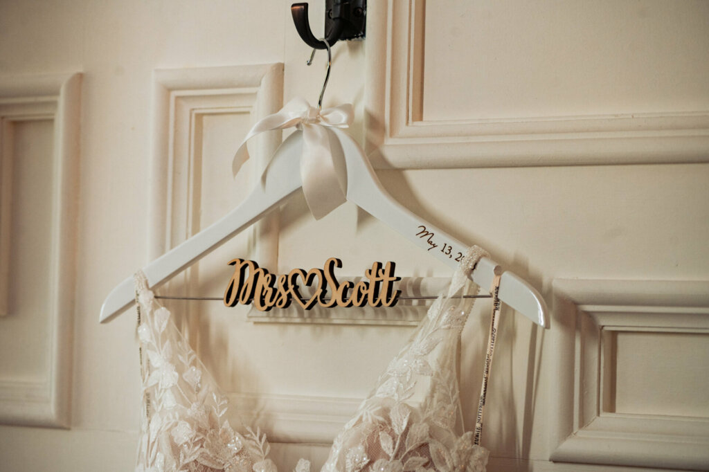 A romantic wedding portrait focuses on an elegant sleeveless bridal gown hanging solitary on a cabinet door, awaiting its bride. Intricate bead and sequin embroidery details the dress front catching the light. The satin hanger displays the bride's new monogrammed initials.