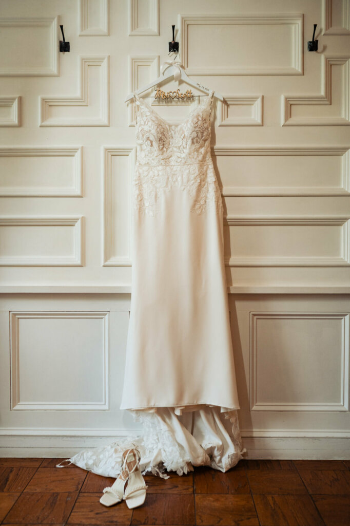 A romantic wedding portrait focuses on an elegant sleeveless bridal gown hanging solitary on a cabinet door, awaiting its bride. Intricate bead and sequin embroidery details the dress front catching the light. The satin hanger displays the bride's new monogrammed initials.