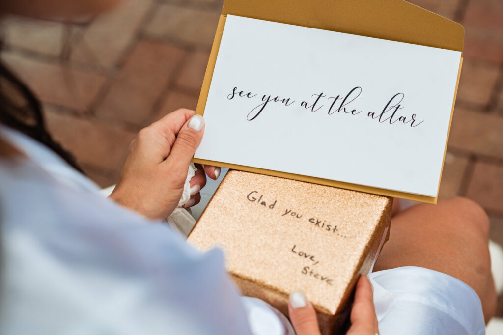 A tender bridal portrait captures a quiet moment of anticipation before a wedding. Seated alone glancing down, an elegantly coiffed bride wearing a silky floral robe clutches a handwritten love letter.