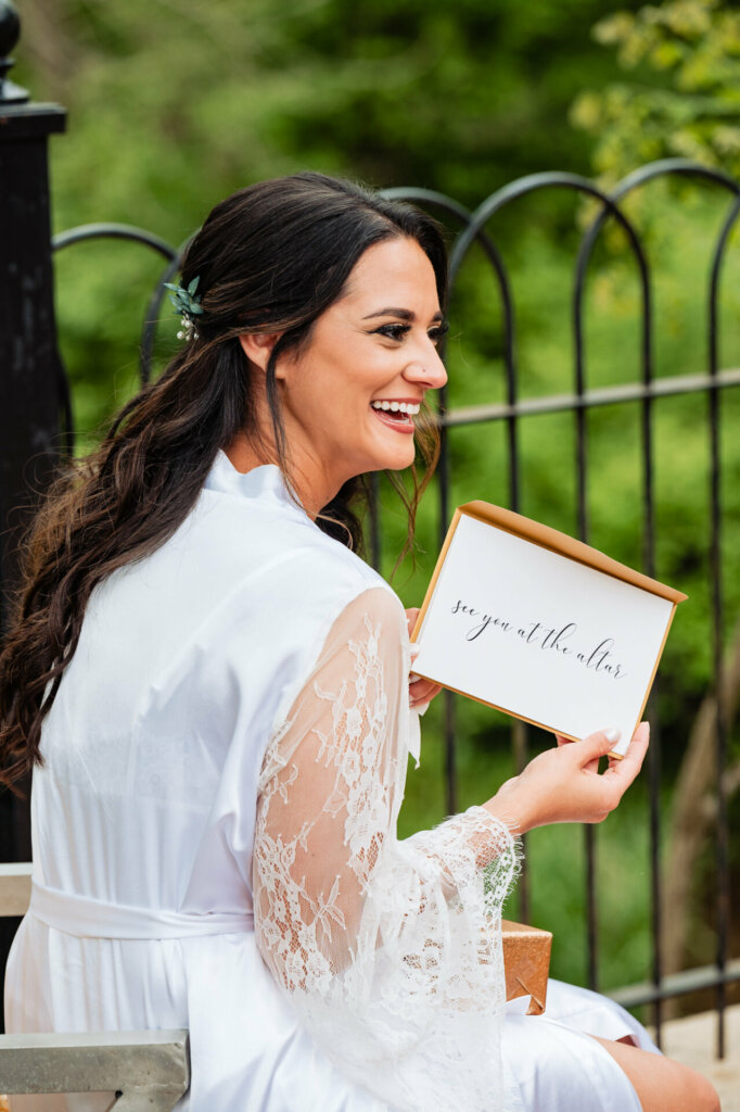A tender bridal portrait captures a quiet moment of anticipation before a wedding. Seated alone glancing down, an elegantly coiffed bride wearing a silky floral robe clutches a handwritten love letter.