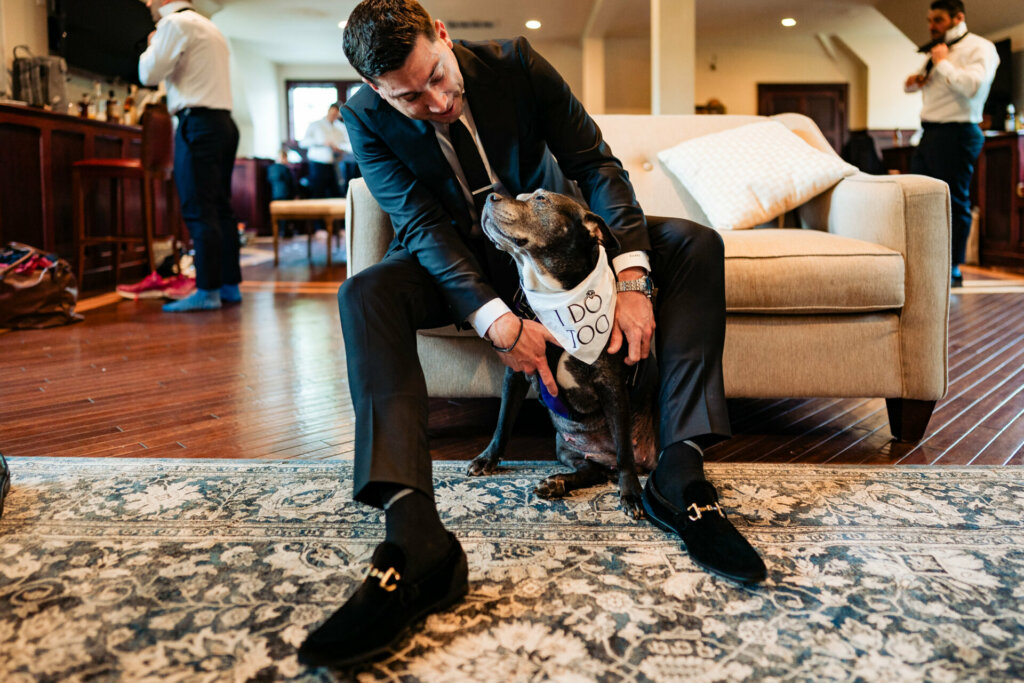 In a playful pre-wedding portrait, a groom-to-be is captured sitting on his couch playing affectionately with his small brown dog.