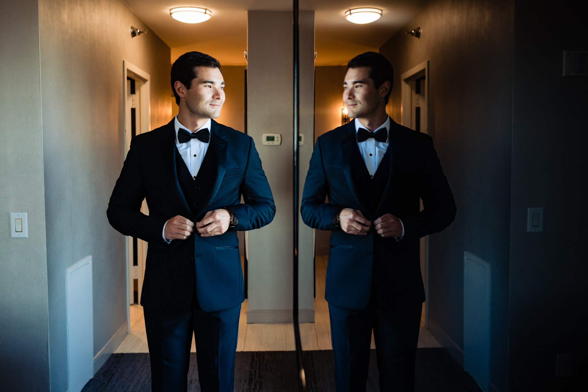 In a moment of quiet contemplation, the groom gazes at his reflection in the mirror, taking in the sight of himself dressed in his wedding attire.