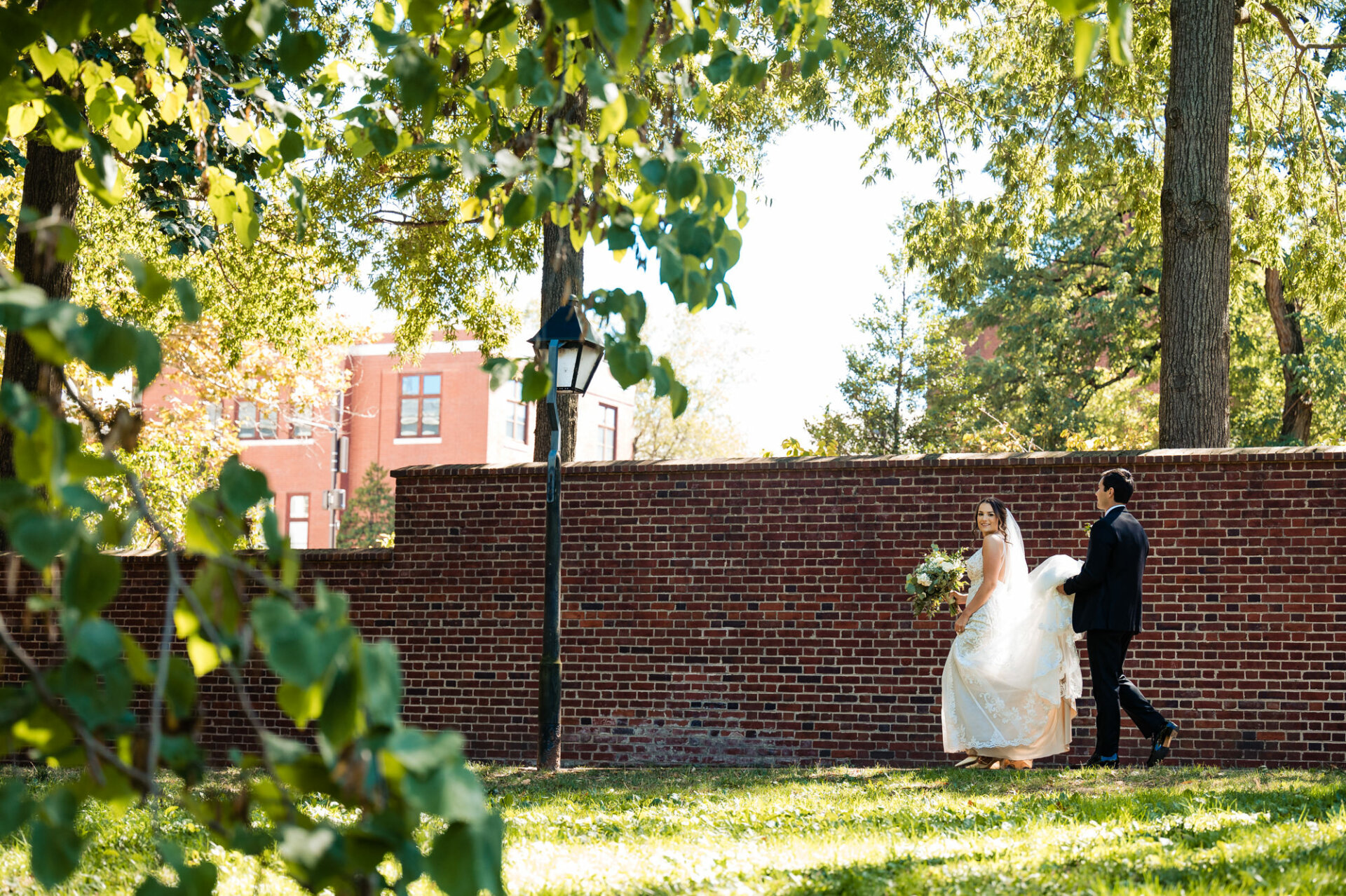 the groom holds the train of his bride's gown as they make their way to their wedding ceremony