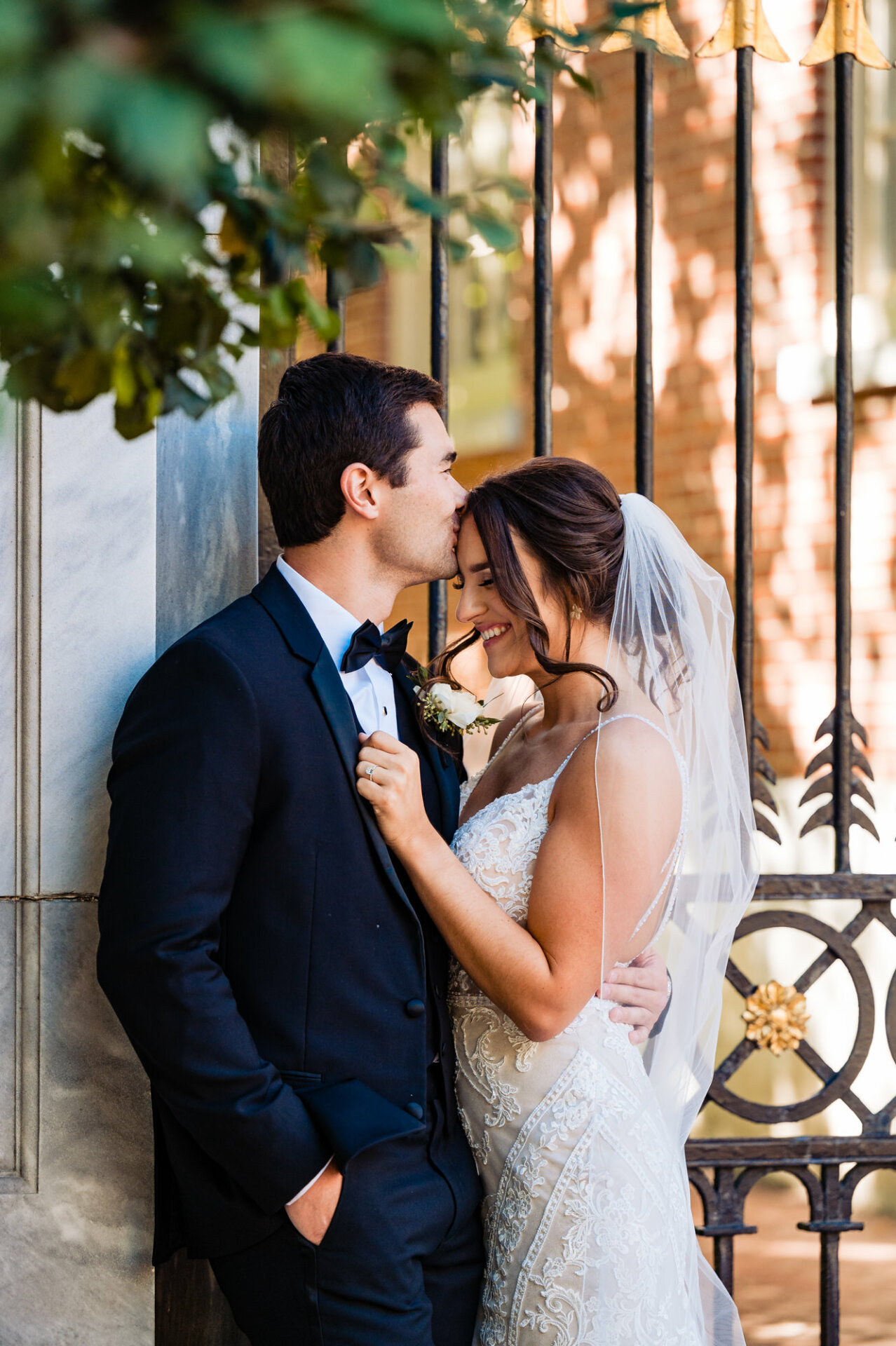 In a captivating moment captured against a backdrop of an elegant steel gate, the bride gets a tender kiss on her forehead from the groom.