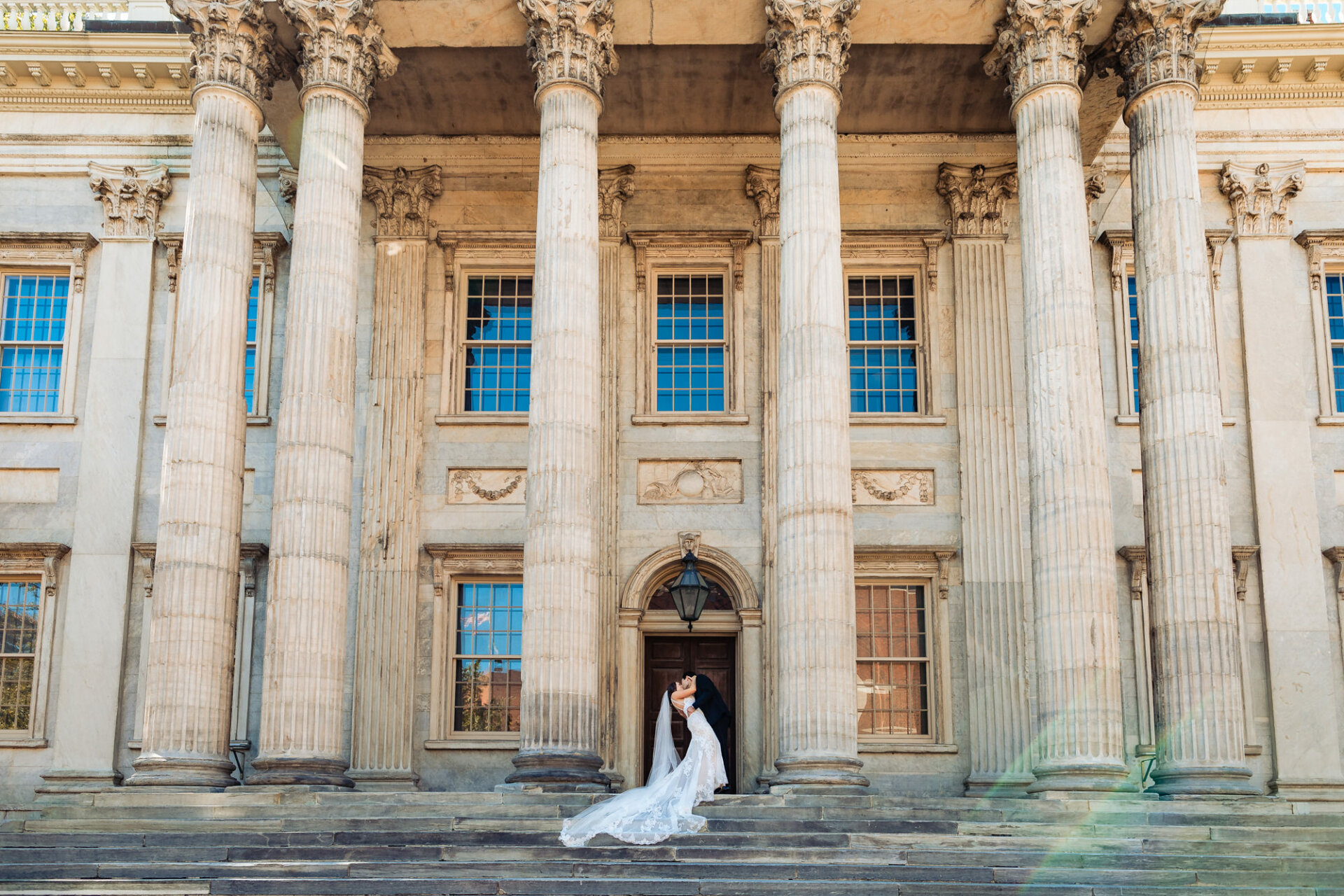 the groom plants a deep and tender kiss on his bride's lips against the towering columns of the First Bank of the United States