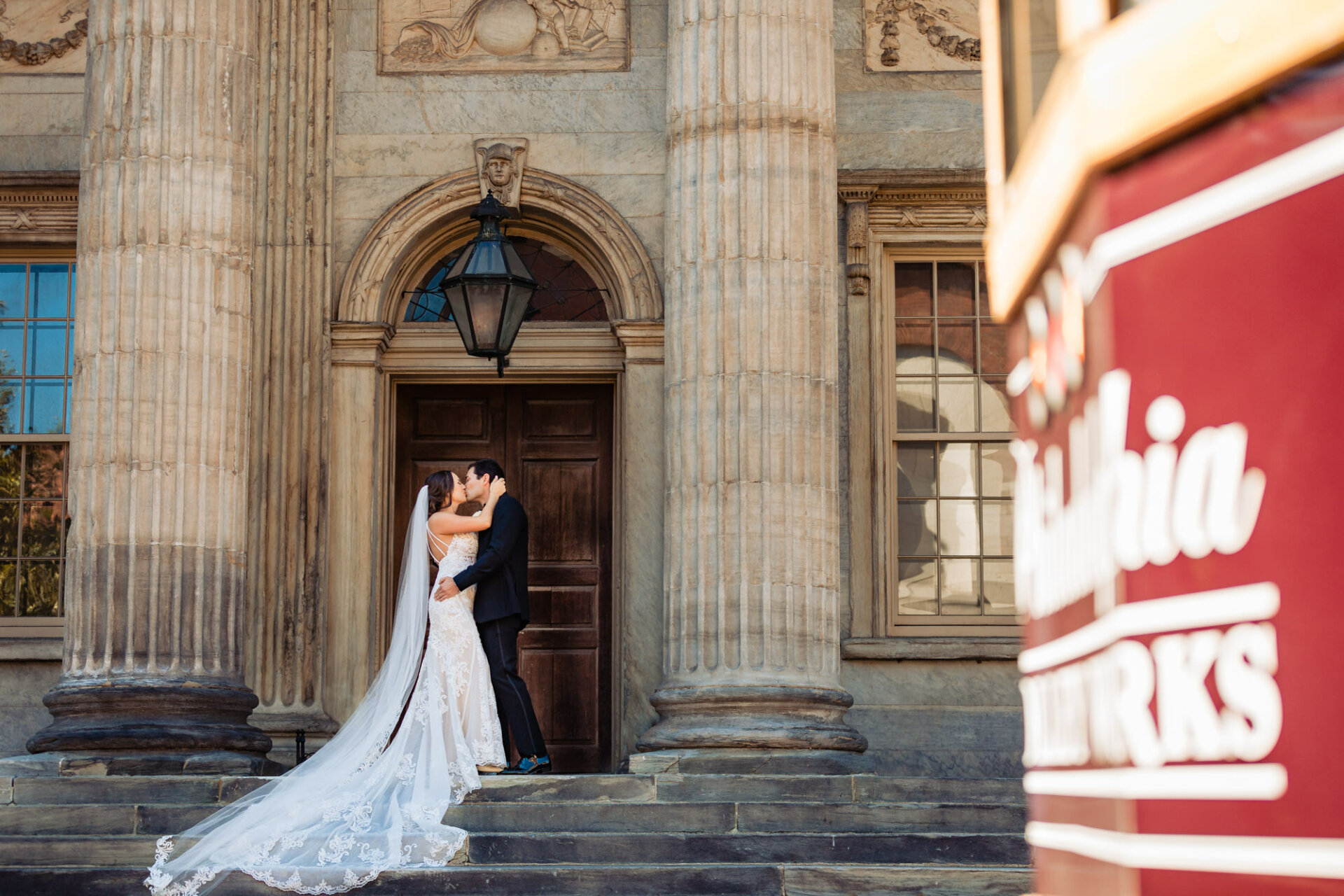 Against the backdrop of towering columns at the First Bank of the United States, the groom tenderly plants a deep kiss on his bride's lips. The grandeur of the architectural marvel amplifies the intensity of their love, casting a majestic aura around the couple.